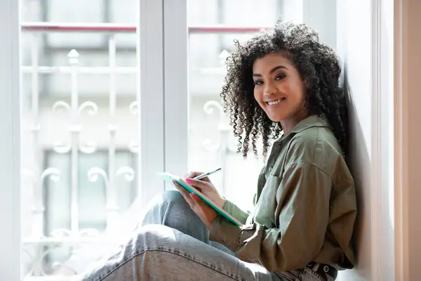 Happy black college student lady sitting by the window at home, holding notebook and taking notes, enjoys weekend of learning and planning indoors, smiling to camera. Concept of creative writing