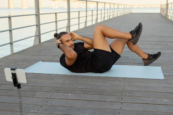 身穿运动服的欧洲运动员在码头上做腹部运动 并通过电话流淌和拍摄他的户外海滨锻炼 健康博客与福利概念 — 图库照片