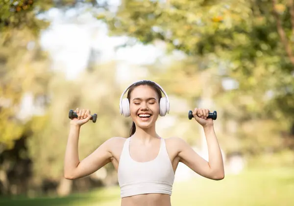 A spirited glad european young woman athlete enjoys her workout, music in a sunny park, lifting dumbbells with headphones on, embodying health and vitality, outdoors in city