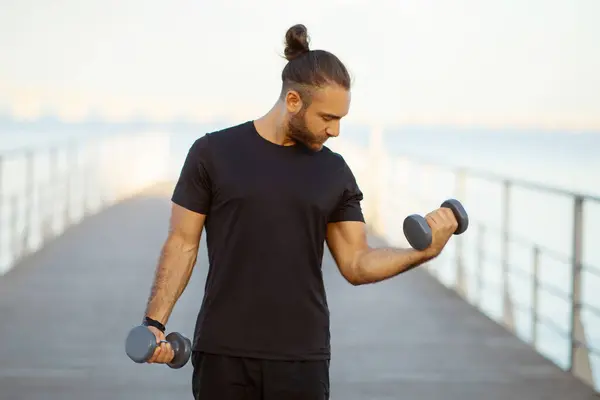 强壮而有进取心的健美运动员在室外用哑铃锻炼他的二头肌 年轻的运动员在海滨码头边的夏季锻炼中促进健康 — 图库照片