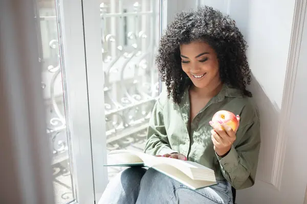 Joy of reading. Happy millennial Brazilian woman indulges in a bestseller book, reads by the window holding apple fruit, enjoying her relaxed weekend in cozy home. Copy space