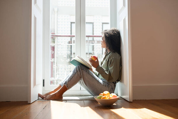 Modern reader. Brazilian lady engrossed in a book reading on floor in comfortable apartment indoor, eating fruits and learning literature by the window at home. Side view shot