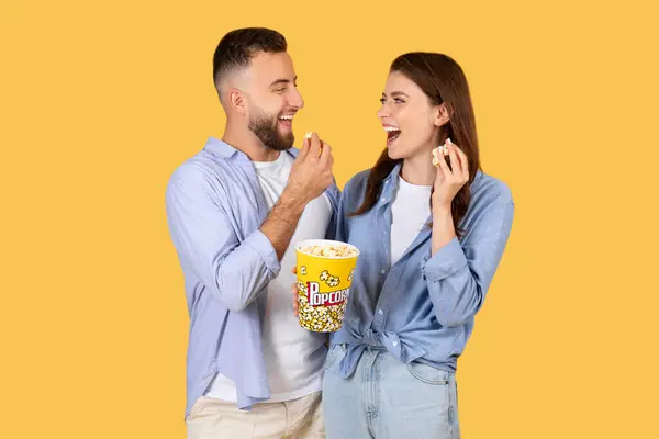 快乐的年轻夫妇共享着轻松的时光 一边一起欢笑 一边吃着五彩缤纷的桶里的爆米花 体现着欢乐与融合 黄色的背景 — 图库照片