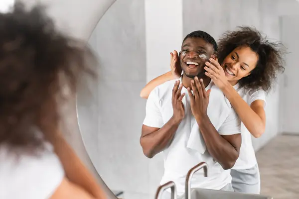 stock image African American loving couple indulges in shared skincare routine at bathroom, as wife applies moisturizer to husbands skin. Love and connection between millennial pair, family pampering