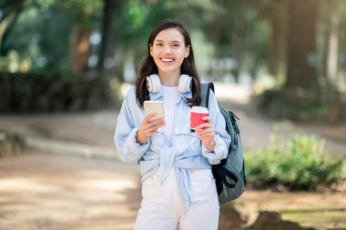 Sıcak bir gülümsemesi olan neşeli beyaz bir öğrenci, elinde akıllı bir telefon ve kırmızı bir fincanla, şehir parkında, açık havada boynunda kulaklıklarla parkta yürümekten zevk alıyor.