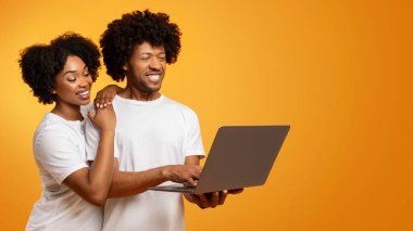 Bin yıllık Afro-Amerikan erkek ve kadın çifti beyaz tişörtler giyip turuncu stüdyo arka planında dizüstü bilgisayar kullanıyorlar, web sörfü yapıyorlar, film izliyorlar, kopyalama alanı olan panorama izliyorlar.