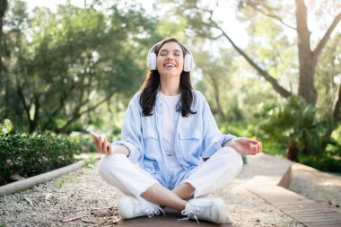 Huzurlu, mutlu, beyaz bir kadın güneşli bir parkta meditasyondan, kulaklıkları takılı ve akıllı telefonlardan keyif alıyor. Huzur ve modern rahatlama yayıyor, boş zamanlarının tadını çıkarıyor.
