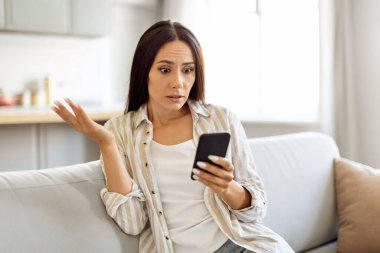 Evde koltukta otururken akıllı telefondan kötü mesaj okuyan genç kadın, elleri cep telefonlu bir kadının nahoş haberlere duygusal tepki göstermesine şaşırdı, Boşluğu Kopyala