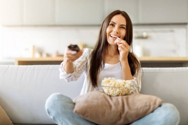 Neşeli genç Avrupalı kadın patlamış mısır yiyor ve elinde uzaktan kumandayla evde televizyon seyrediyor, mutlu bir milenyum kadını rahat bir koltukta oturuyor, hafta sonu tatilinin tadını çıkarıyor.