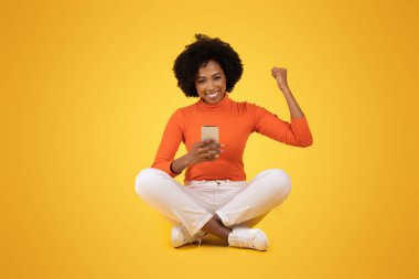 Doğal kıvırcık saçlı neşeli bir Afro-Amerikan kadını, sarı arka planda bağdaş kurmuş bir şekilde cep telefonunu tutarak zaferini kutluyor.