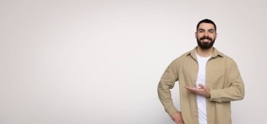 Eli yanda olan neşeli sakallı bir adam, yazı veya ürün için boş bir alan gösteriyor, geniş bir arkaplanda bej bir gömlek ve beyaz bir tişört giyiyor.