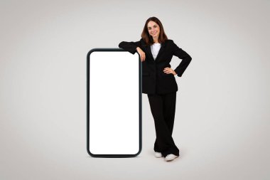 Bin yıllık kendine güvenen, özel dikim takım elbiseli beyaz kadın boş bir ekranla dev bir akıllı telefona yaslanıyor ve teknolojiden anlayan iş çözümleri kavramını gözler önüne seriyor.