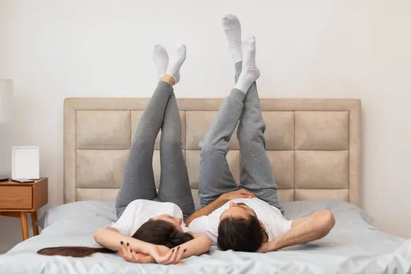 Couple enjoys playful moment in cozy bedroom, lying on their backs on the bed with legs up in the air, dressed in casual home wear, creating relaxed and cheerful atmosphere