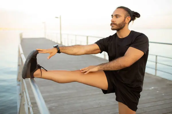 アクティブストレッチ ブラックフィットウェアの男性は 屋外の桟橋に立って脚を伸ばし 海辺の背景で柔軟性と健康的なライフスタイルを促進します アクションコンセプトにおけるフィットネスとウェルネス — ストック写真