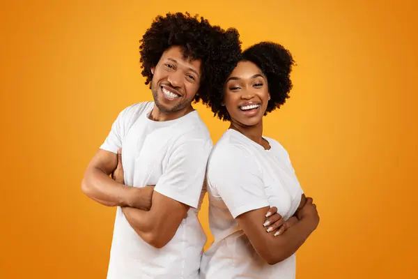 Positive millennial black man and woman wearing white t-shirts standing back to back holding arms crossed on chest, smiling at camera, posing on orange background. Friendship, teamwork