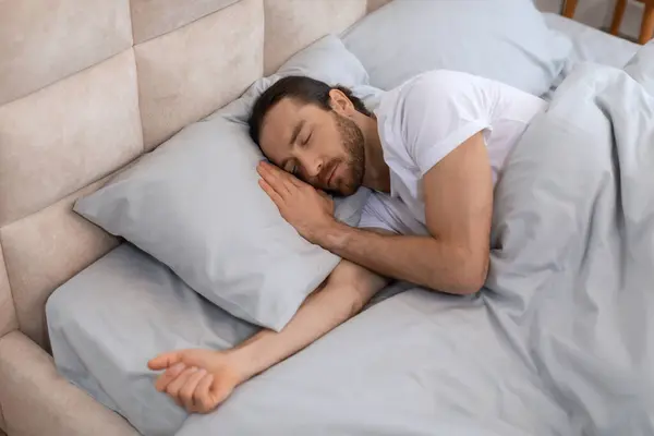 Man in white t-shirt is sound asleep, showcasing the tranquility of resting in cozy bed with plush pillows and silky sheets, evoking serenity, above view