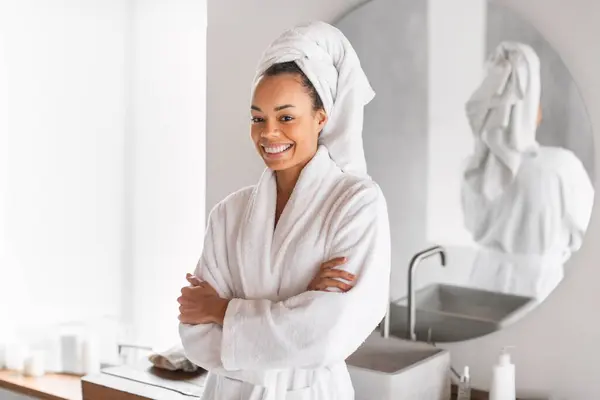 Daily beauty routine. African American millennial lady standing with folded arms in bathrobe, enjoys her morning self care rituals, smiling to camera in modern bathroom. Pampering and wellness