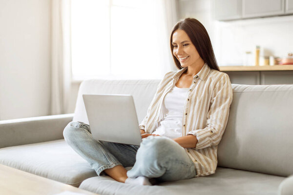 Расслабленная молодая женщина наслаждается работой из дома, комфортно используя ноутбук, сидя на диване в уютной гостиной, счастливая женщина тысячелетия делает онлайн-работу, копировать пространство