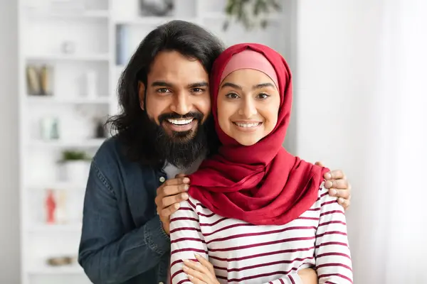 Portrait Of Loving Muslim Couple Posing Together At Home, Young Arabic Spouses Embracing And Smiling At Camera, Man And Woman In Hijab Bonding While Posing In Living Room Interior, Copy Space