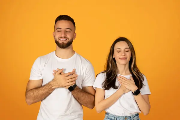 充满感激之情的年轻的高加索夫妇 紧闭双眼 手拉手 在温暖的橙色背景下 表达着诚挚的情感和感激之情 身穿白色T恤和牛仔裤 — 图库照片