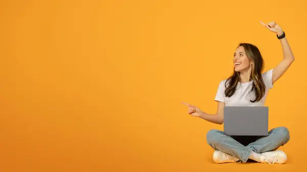 身穿白色T恤和蓝色牛仔裤的快乐的高加索女人 双腿交叉坐在地板上 用笔记本电脑向上举着手势 带着愉快的表情面对着充满活力的橙色背景 — 图库照片