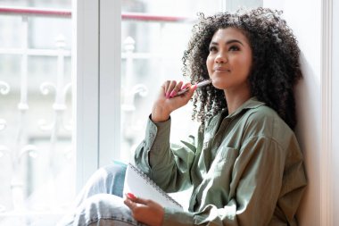 Afro-Amerikalı iş kadını, rahat oturma odasındaki pencerenin kenarına not defteriyle oturmuş yazmayı ve düşünmeyi planlayıp yaratıcı bir şekilde yazmayı seviyor. Boş boşluk
