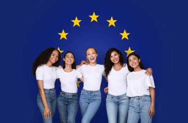 Beyaz bluzlu ve kot pantolonlu beş genç kadın, Avrupa Birliği bayrağına sarılıp gülümsüyorlar, arkadaşlığı ve birliği temsil ediyorlar, stüdyo. Güzellik, sağlık hizmetleri