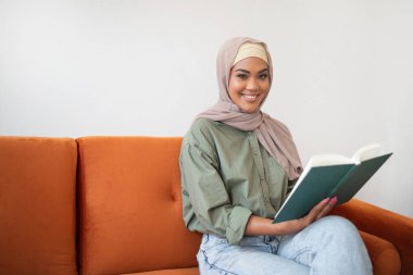 Brezilya 'lı başörtüsü takmış kadın elinde kitapla kanepeye oturuyor, boş zamanlarını okumaya dalmış, evdeki modern oturma odasında kameraya gülümsüyor, hem konfor hem de eğitimi kucaklıyor.