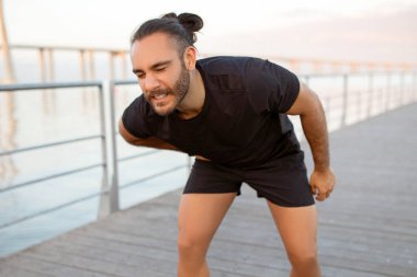 Sportif adam dışarıda idmandan sonra acı içinde kıvranıyor, egzersizin gerginliğini hissediyor, nefes nefese, koşucu yan dikiş atmak için duruyor, yüzü rahatsızlık içinde kıvranıyor.