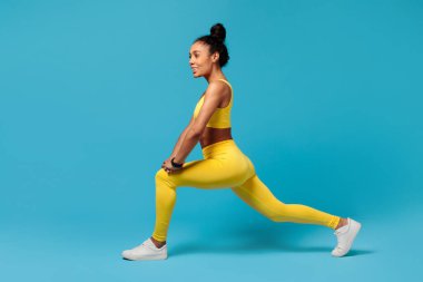 Aktif yaşam tarzı, egzersiz. Sarı spor kıyafetli, atlama, esneme ve güçlendirme bacaklı Milenyum siyahi sporcusu, stüdyoda mavi arka planda egzersiz yapıyor. Spor motivasyonu