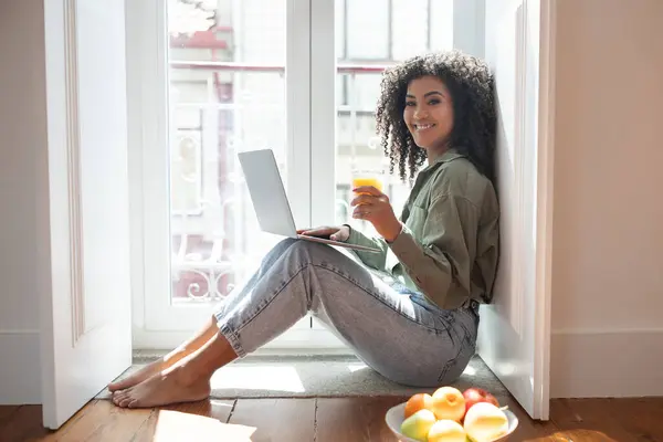 Brazilian freelancer lady sitting with laptop and embraces convenience of working from home, enjoying refreshing juice drink in front of computer indoor. Distance online education and business