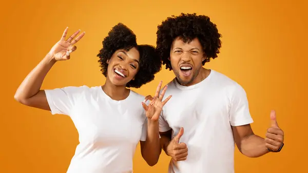 Emotional stylish african american couple gesturing on orange studio background. Happy millennial black man and woman wearing white t-shirts showing thumb ups and okay gestures, panorama