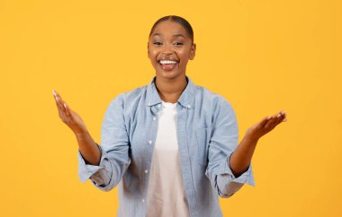 Kameraya gülümseyen ve sarı arka planda el kol hareketi yapan neşeli siyahi kadının portresi. Stüdyo portresi. Ticari tanıtım kavramı