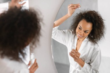 Modern banyodaki aynanın yanındaki dalgalı saçlarına krem serumu sürerek mutlu siyah kadın saç bakım rutini yapıyor. Sağlık, doğal güzellik ürünleri. Seçici odak