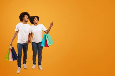 Heyecanlı genç siyah çift birlikte alışveriş yapıyor, reklam için boş yeri işaret ediyorlar. Mutlu Afro-Amerikan erkek ve kadın ellerinde renkli kağıt poşetler, turuncu stüdyo geçmişi var.
