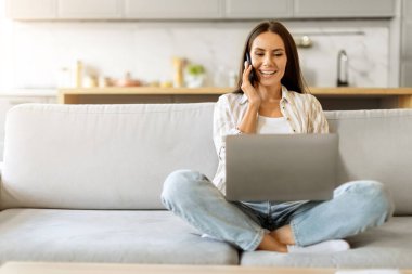 Uzaktan kumanda. Telefonla konuşan ve dizüstü bilgisayar kullanan gülümseyen genç bir kadın oturma odasında oturan, bilgisayar üzerinde çalışan ve cep telefonu kullanabilen güzel bir Avrupalı kadın,