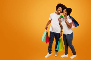 Mutlu Afro-Amerikan çifti birlikte alışveriş yapıyor, reklam için boş alanı kopyalıyor. Neşeli siyah adam ve kadın ellerinde renkli kağıt poşetler, turuncu stüdyo arka planı, tam boy.