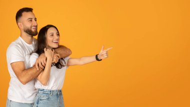 Beyaz tişörtlü ve kot pantolonlu mutlu beyaz bir çift, kadın arkasında bir adam, şakacı bir şekilde yan gösteren, hem gülümseyen ve uzak bakan parlak turuncu bir arka plan