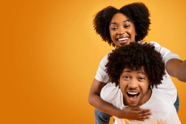 Milenyum Afro-Amerikan çifti turuncu stüdyo arka planında selfie çekiyor. Sevimli siyah bayan erkek arkadaşını sırtında taşıyor ve elinde kamera, boş fotokopi alanı.