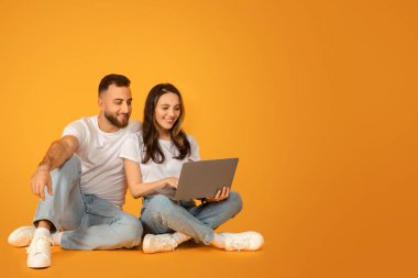 Beyaz bluzlu ve kot pantolonlu rahat ve mutlu beyaz bir çift yerde bağdaş kurmuş, dizüstü bilgisayarlarını paylaşıyorlar turuncu bir arka planda, stüdyoda, tam boy