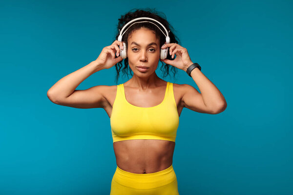Fitness vibe. Motivated African American woman in yellow activewear stands confidently in studio, putting on wireless earphones, ready for healthy workout while listening to music on blue backdrop