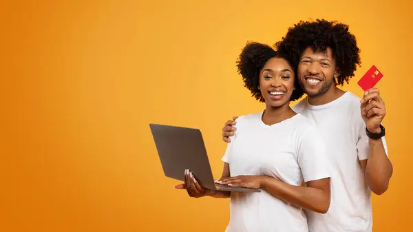 Happy young black man and woman wearing white t-shirts banking or shopping online isolated on orange studio background, holding laptop computer and credit card, panorama with copy space