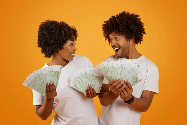 Cashback, lottery win, Emotional happy millennial african american spouses showing money euro banknotes and looking at each other, isolated on orange studio background