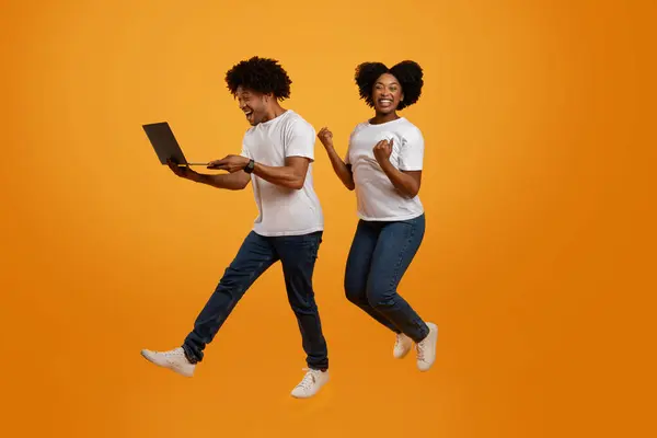 Emotional happy millennial african american man and woman spouses with laptop computer jumping up in the air and gesturing over orange studio background, got cashback, win lottery, full length