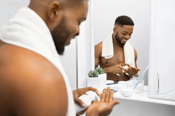 stock image Smiling afro man with naked torso squeezing cream on his hand, reflecting in mirror at bathroom. Mans skin care concept