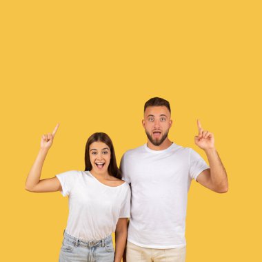 Beyaz tişörtlü heyecanlı genç çift şaşırmış ve mutlu bir ifadeyle yukarıya doğru bakıyor, sarı arka planda ilginç bir şeye dikkat çekiyorlar.