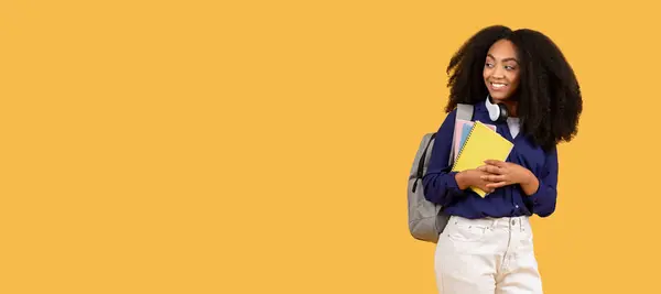 快乐的年轻黑人女人 带着耳机 拿着笔记本 微笑着看了看空旷的空间 站在温暖的黄色背景上 全景尽收眼底 — 图库照片