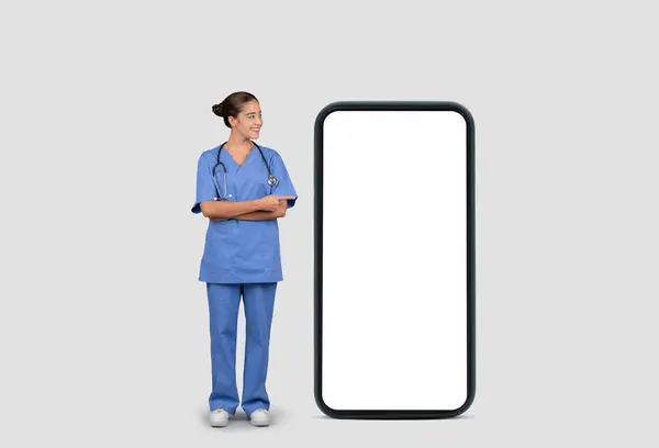 Uśmiechnięta Europejska Pielęgniarka Niebieskim Mundurze Stetoskopie Stojąca Obok Gigantycznego Smartfona — Zdjęcie stockowe