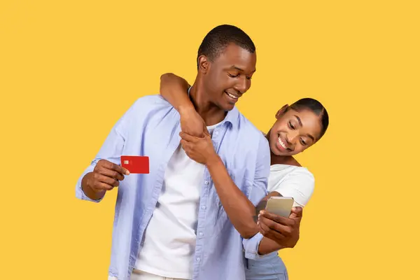 黑人夫妇上网购物 男人拿着信用卡 女人靠在他的肩上用智能手机 在黄色背景下分享快乐时光 — 图库照片