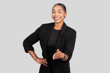 Afro-Amerikalı bir iş kadınının elini sıkması, parlak bir gülümseme, anlaşma imzası, temiz gri bir arka planda şık siyah bir takım elbise giymesi.